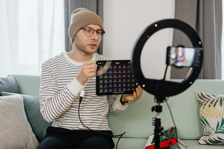 Adult man wearing a beanie records a tech review vlog indoors using a smartphone and ring light.